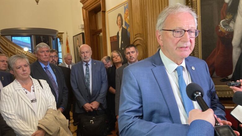 A man with grey hair and glasses, wearing a blue suit, white collared shirt and blue tie, speaks into several reporters' microphones as a number of other people behind him look on.