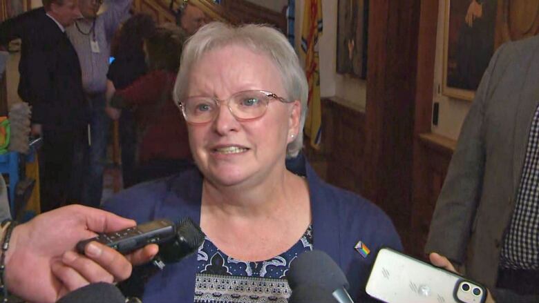 A woman with blond hair and glasses, wearing a royal blue jacket, speaks into several reporters' microphones and tape recorders.