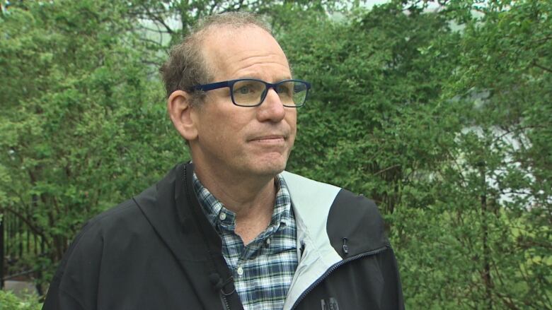 A white man with glasses and a black jacket stands in a backyard surrounded by trees