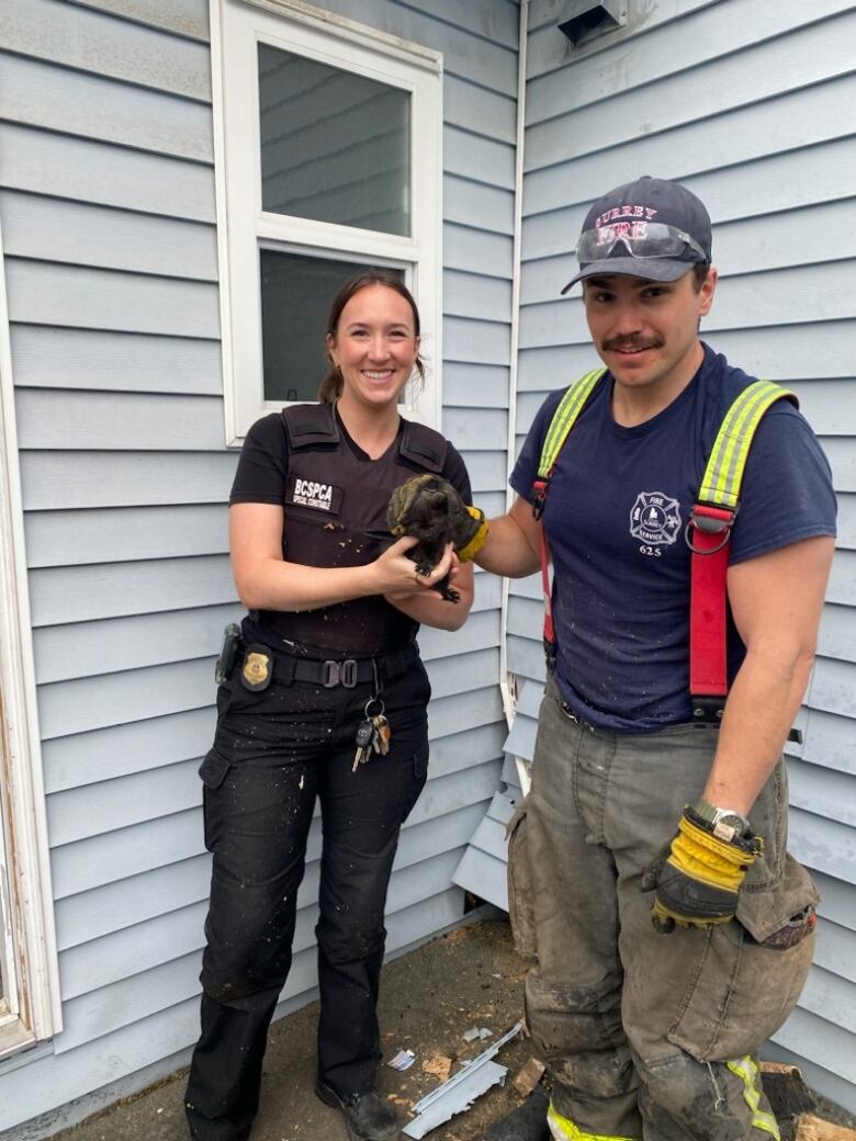 A woman wearing a BC SPCA vest and a firefighter stand holding a tiny black kitten outside a home with a visible hole cut in the side.