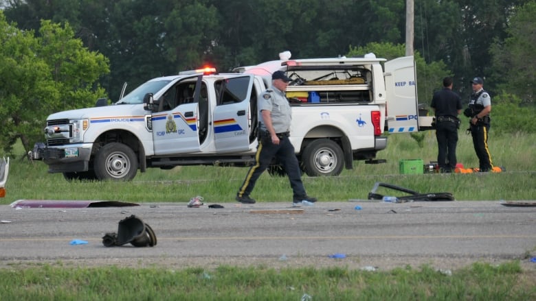 A police officer is seen walking on a road following a collision. Vehicle debris is pictured all over the road. A police truck is seen in the background with its doors open, and two officers standing at the back of it talking.