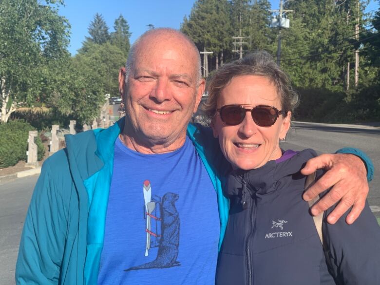 A man in a blue shirt and jacket smiling next to a woman in a black jacket and sunglasses with his arm around her.