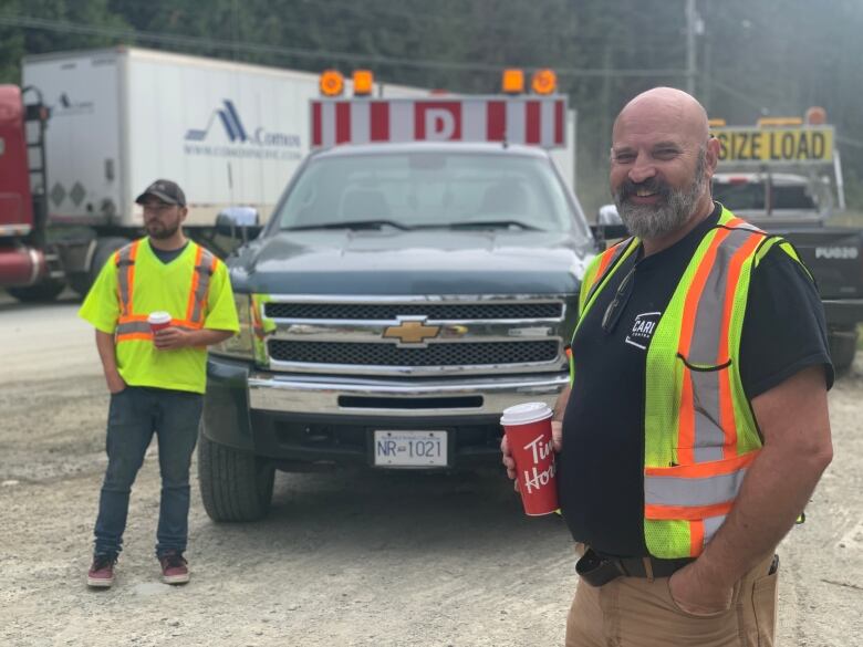 A man with a safety vest and a tim hortons cup smiles at the camera, he is standing in front of a few trucks with 