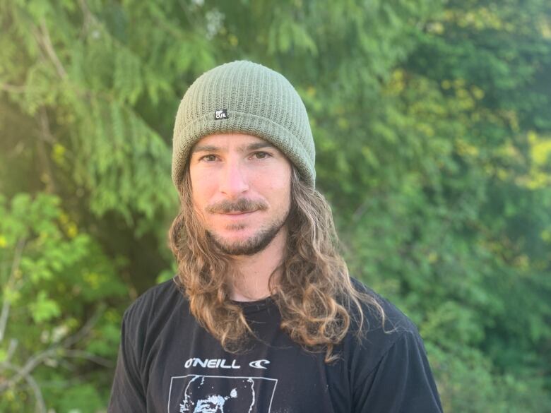 A man with long brown hair and a green toque stands in front of a tree-lined area.