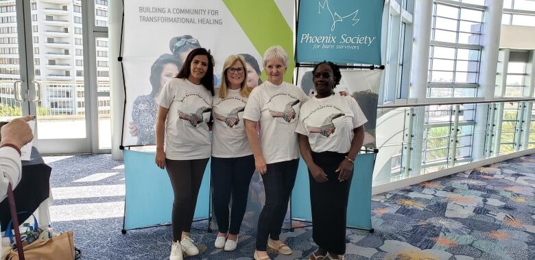 Four women in t-shirts smile at the camera in front of banners that say Phoenix Society