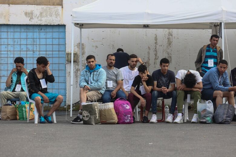 Eleven men sit, looking dejected and tired, in front of a concrete building, a collection of overstuffed shopping bags at their feet. 