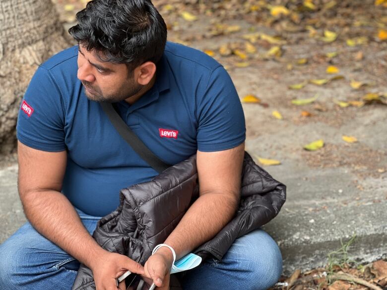 A heavyset man with dark hair wearing a dark blue polo shirt and jeans sits on a curb holding a jacket and a phone.  