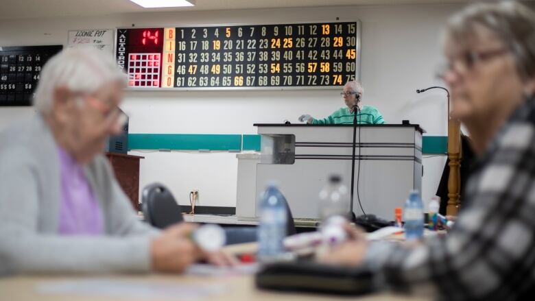 Two people sit at a table playing bingo. A person stands in front of a microphone in the background.