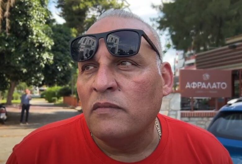 A man in a red t-shirt with short, greying hair wears sunglasses propped up on his forehead as he speaks to members of the media. 