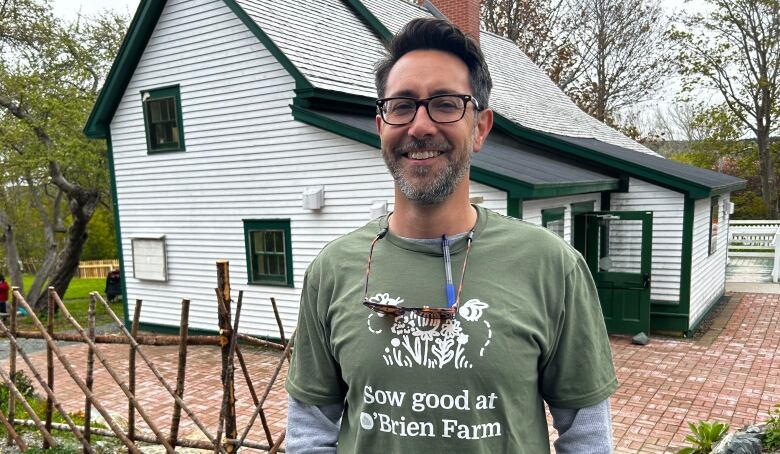 A man with glasses wearing a green t-shirt standing in front of a white house. 