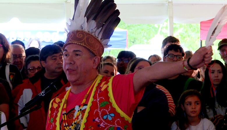 A Wolastoqey First Nations chief in a red short-sleeve shirt and red traditional garment speaks into a microphone to address a crowd. His left arm is extended straight out to his left and he holds a feather in that hand.