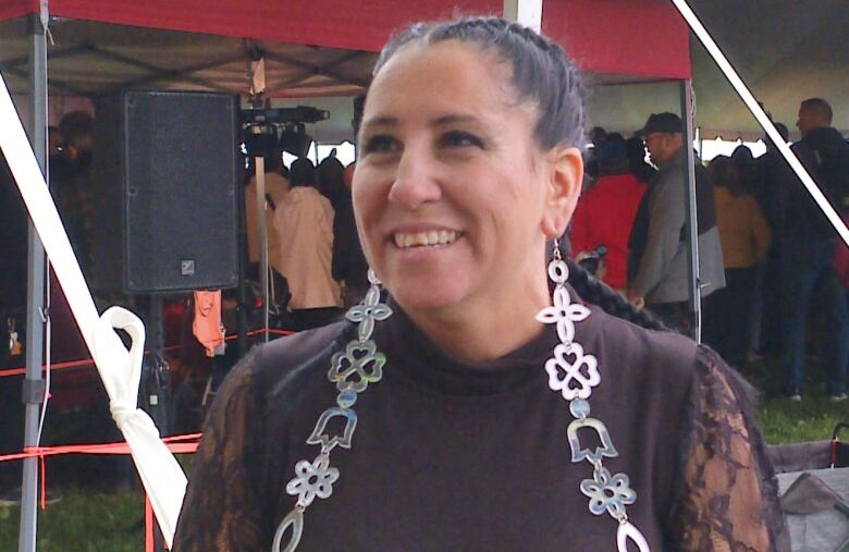 A woman with long dark hair in tight braids behind her, smiles slightly to the left of the frame. She is wearing a black long sleeve blouse with sheer sleeves and long silver earrings with large symbols that extend past her chest.