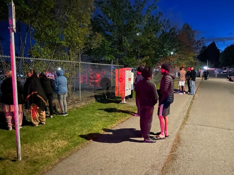 Onlookers gather and look through a fence where police sirens cast a red and blue glow.