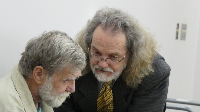 An older man with long grey/brown hair and a grey beard leans down to speak with another older man with grey hair and a white beard.