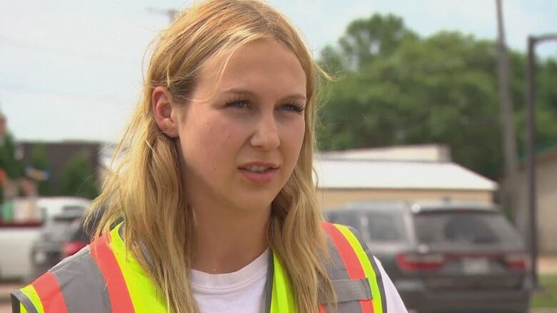 A teen girl with medium-length blonde hair, wearing a neon safety vest, is pictured looking forward.