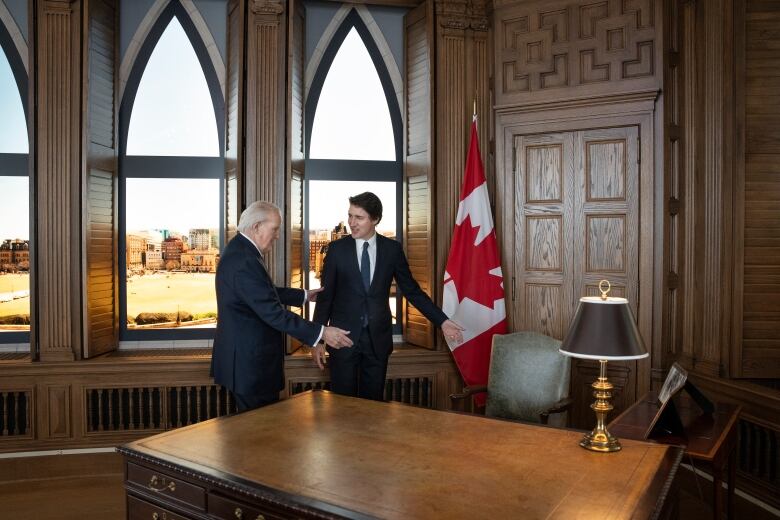 Two men, one middle-age and one older, both wearing suits, stand in a wood-clad room next to a Canada flag and a desk.