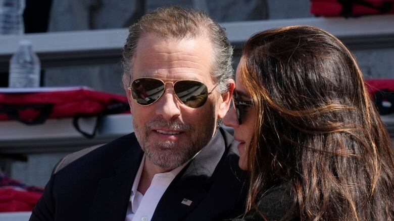 A man and a woman in sunglasses are shown speaking outside.