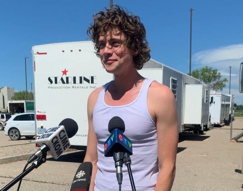 A man with blonde curly hair is wearing a white tank top and standing in front of mics being interviewed. 