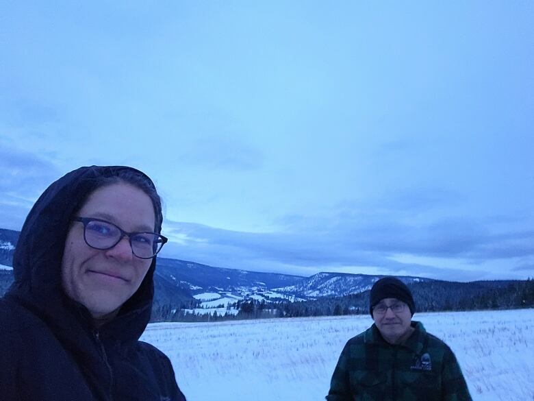 A man and a woman are pictured in a selfie photo on a snow-covered field with mountains in the background. 