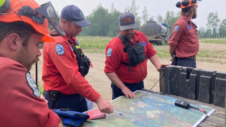 Four men in red, long-sleeved, button-down shirts, and navy blue baseball caps stand around the back of a pickup truck and look at a map.