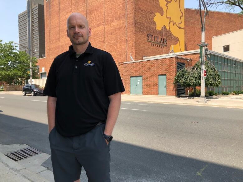 A man in a black t-shirt stands in front of a St. Clair College building