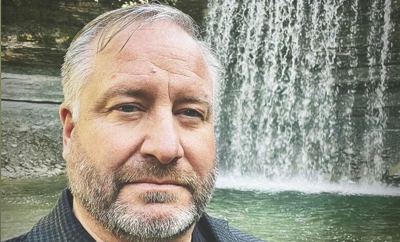 A man with a grey beard stands in front of a waterfall 