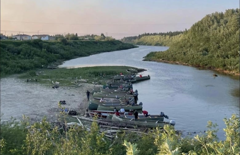 A row of boats is parked on a beach on a winding river, with houses up on a hill 