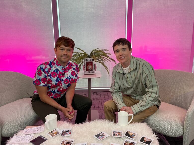 Actor and author Elliot Page is seated in a chair next to with Here & Queer host Peter Knegt as they pose for the camera.