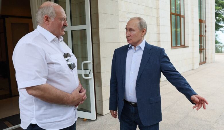 Two older balding men, one wearing a white short-sleeved shirt and the other wearing a blue blazer, speak outside a building.