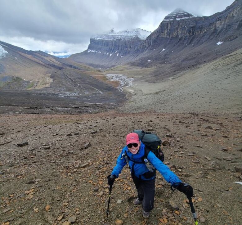 A woman stands in a high altitude pass with hiking poles in her hands and a large backpack on her back.