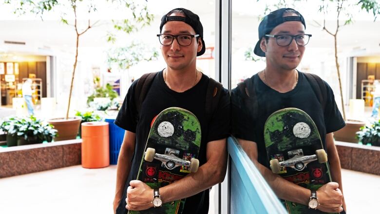 A man holding a skateboard leans against a mirror.