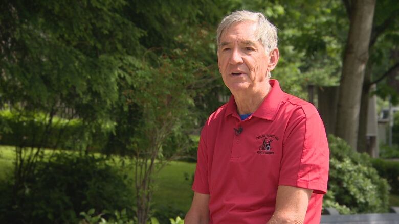 A man with white hair and wearing a pink polo shirt is interviewed on camera