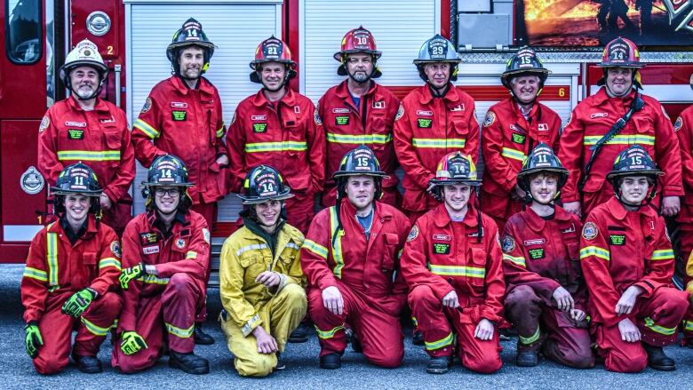 Firefighters in red uniforms and one in yellow and shown in two rows in front of a fire truck and wearing firefighter helmets.