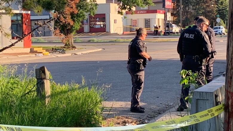 Three police officers in dark uniforms look around urban street cordoned off by yellow tape.