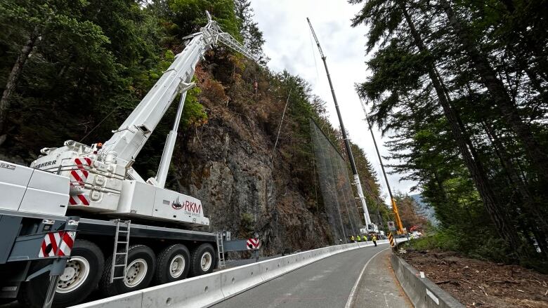 A crane truck helps install protective netting on a rock face beside a closed highway as workers operate on the roadway.