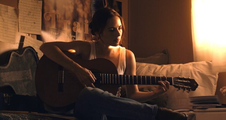 A still from a film shows a sombre woman in a sunlit room playing a guitar.