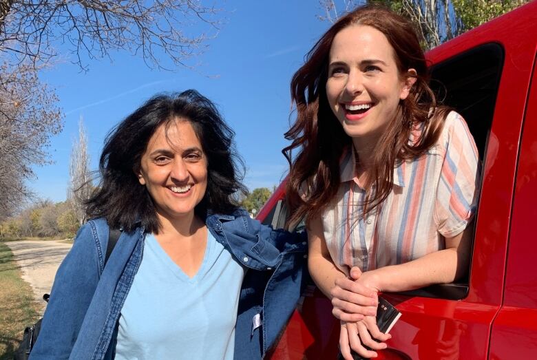 A smiling woman stands beside a red pickup truck, with a woman sitting inside leaning out the driver's-side window, smiling.