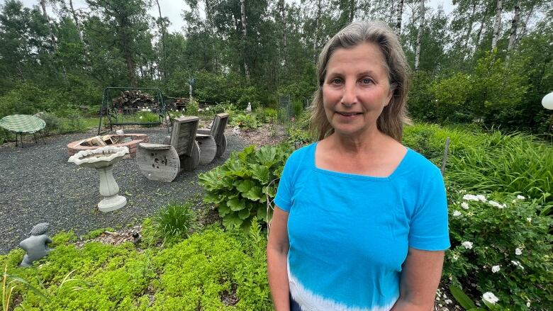 A woman with shoulder-length gray hair wearing a blue shirt stands in front of a garden while speaking to a reporter. 