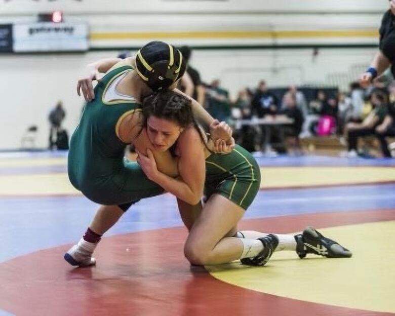 Two women in green leotards participate in a wrestling match inside a school gymnasium.