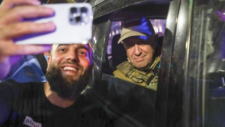 A bearded man smiles as he holds his smartphone up to take a selfie. A man in military garb smiles over his shoulder, seen through the rolled-down window of a vehicle.