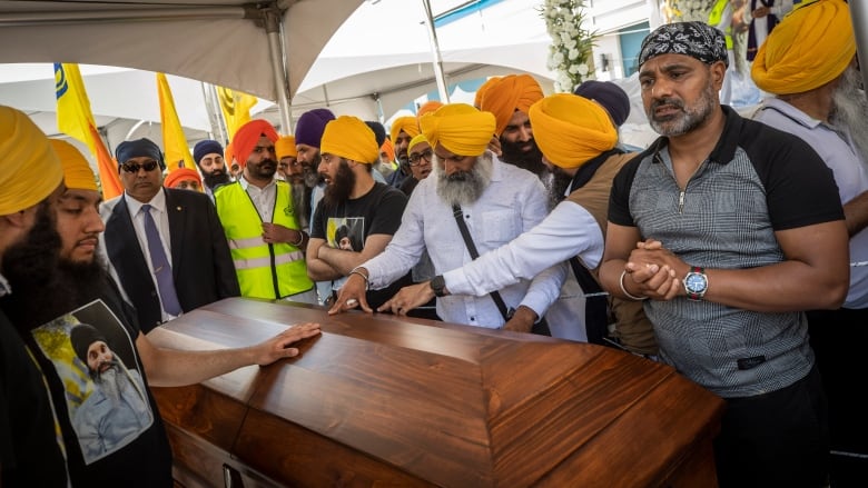 A crowd of men wearing a mix of orange and blue turbans and head coverings place their hands on a wooden casket in the centre.