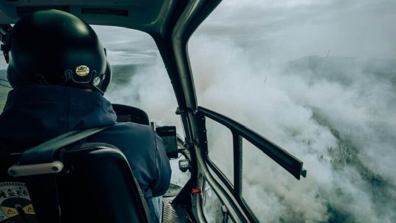 A helicopter pilot is photographed from behind as he flies over wildfires. 