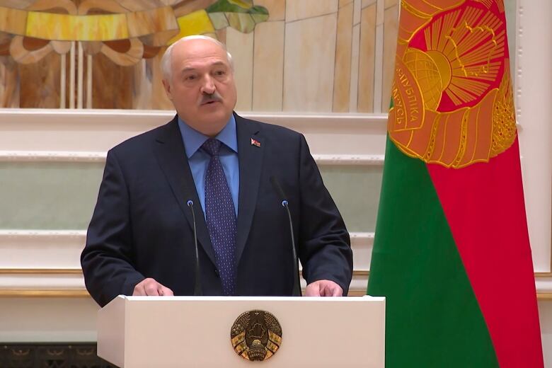 A man in a suit speaks at a podium, with a national flag behind him.