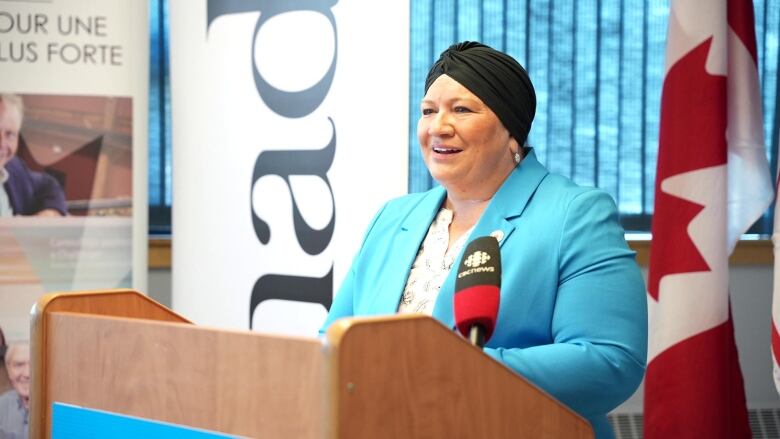 A woman in a blue blazer with a black head wrap stands in front of a podium.