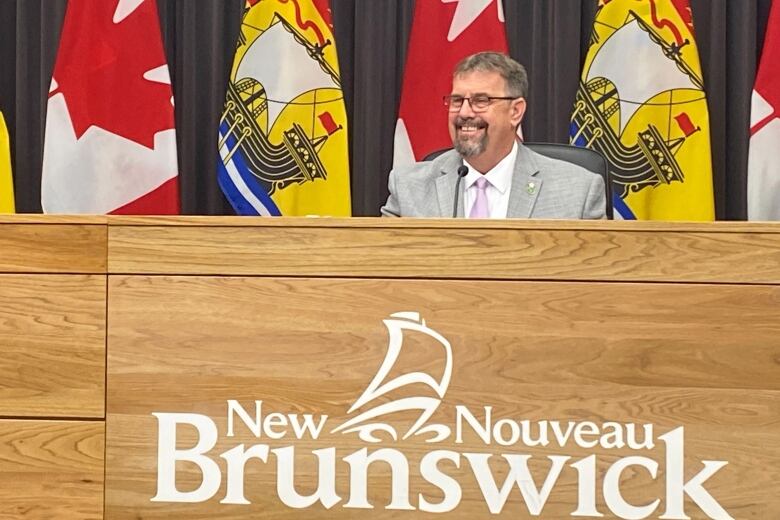 A man with glasses and a goatee and moustache smiles as he sits behind a barrier that says 