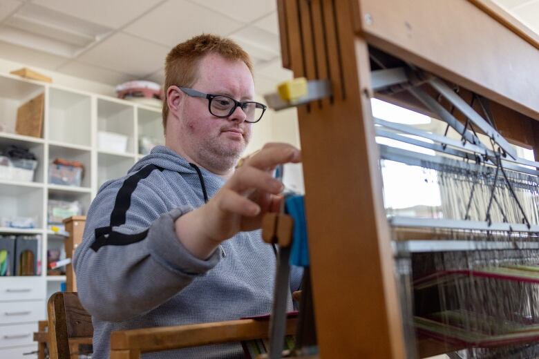 A red-haired person in a grey hoodie works on a weaving loom. 