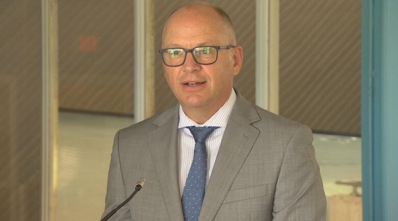 An older man wearing black rimmed glasses and a grey suit is pictured speaking.