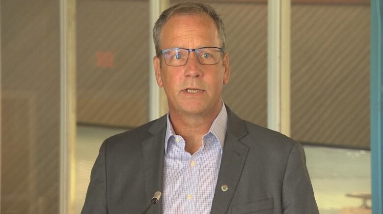 An older man wearing thin, black-rimmed glasses and a grey suit is pictured speaking.