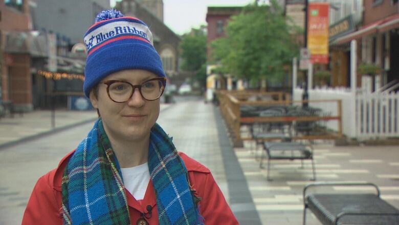 A woman wears a red coat, a blue, white and red hat. She's also wearing glasses and a Nova Scotia tartan scarf.  Behind her are tables, chairs and restaurant patios. 