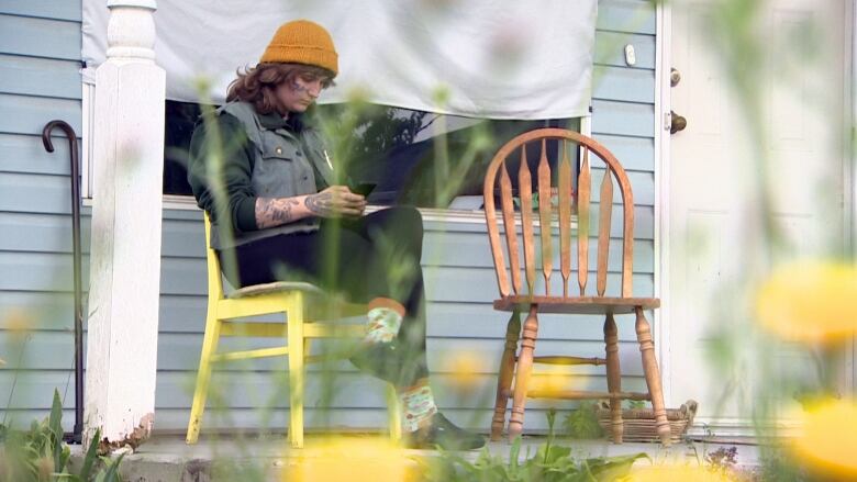 A tattooed person wearing a yellow toque and a denim vest over a long-sleeved shirt sits on a yellow wooden chair in front of a blue-siding-clad home with sheets over the windows.